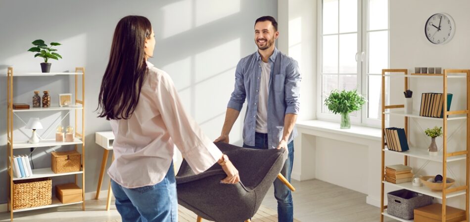 Young,couple,carrying,armchair,into,their,living,room.,smiling,man